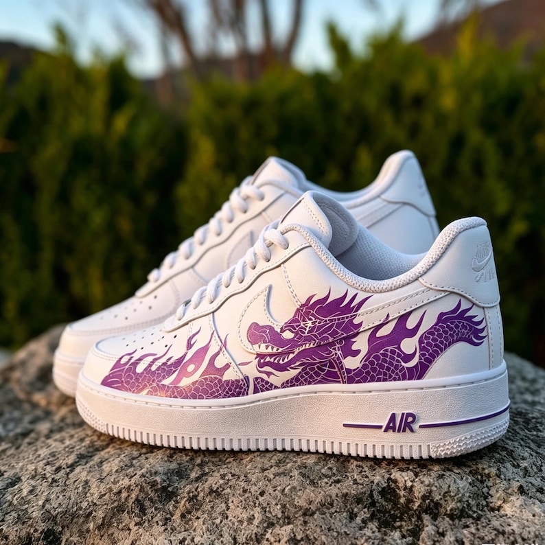 May include: White sneakers with a purple dragon design on the side. The shoes have a classic low-top style with white laces and a white sole. The word "AIR" is printed in purple on the sole.