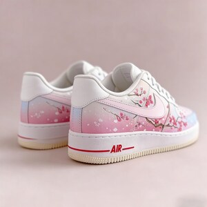 Op de afbeelding: Witte en roze sneakers met een kersenbloesemdesign. De schoenen hebben een verloop van roze en blauwe tinten, met een rood "AIR" detail op de zool. Het ontwerp bevat roze kersenbloesemtakken.