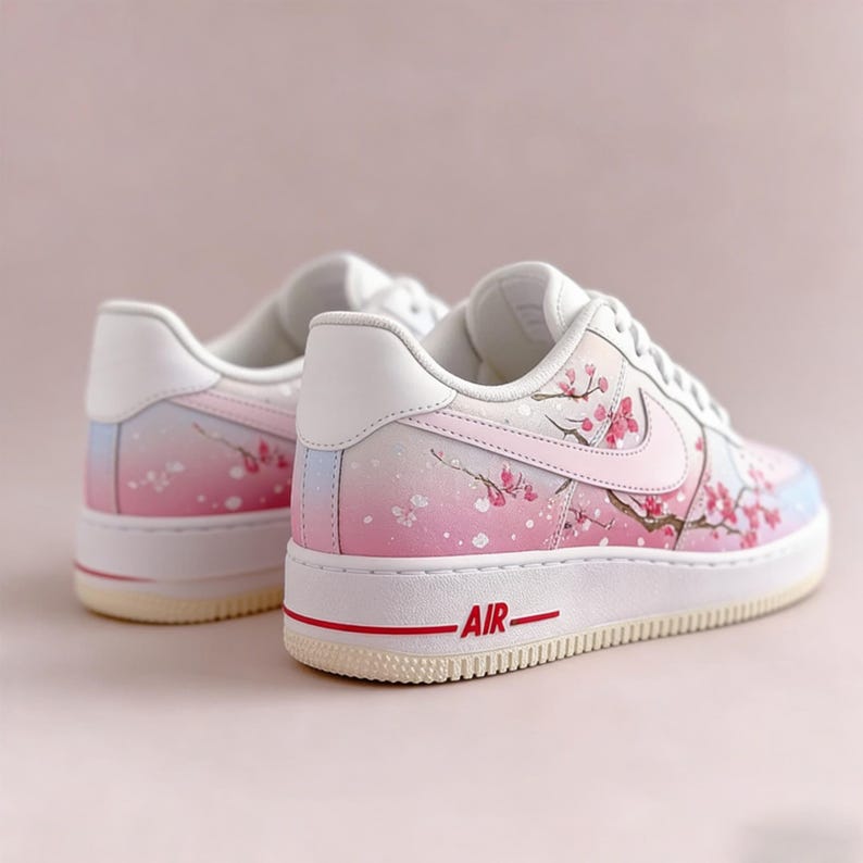 Op de afbeelding: Witte en roze sneakers met een kersenbloesemdesign. De schoenen hebben een verloop van roze en blauwe tinten, met een rood "AIR" detail op de zool. Het ontwerp bevat roze kersenbloesemtakken.