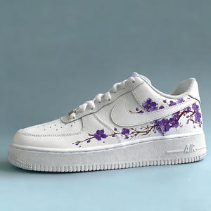 May include: White leather sneaker with a thick sole, featuring a purple floral branch design. The shoe has a classic lace-up closure and the Nike swoosh logo on the side. The sole reads "AIR".