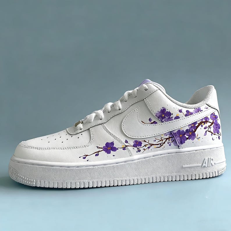 May include: White leather sneaker with a thick sole, featuring a purple floral branch design. The shoe has a classic lace-up closure and the Nike swoosh logo on the side. The sole reads "AIR".