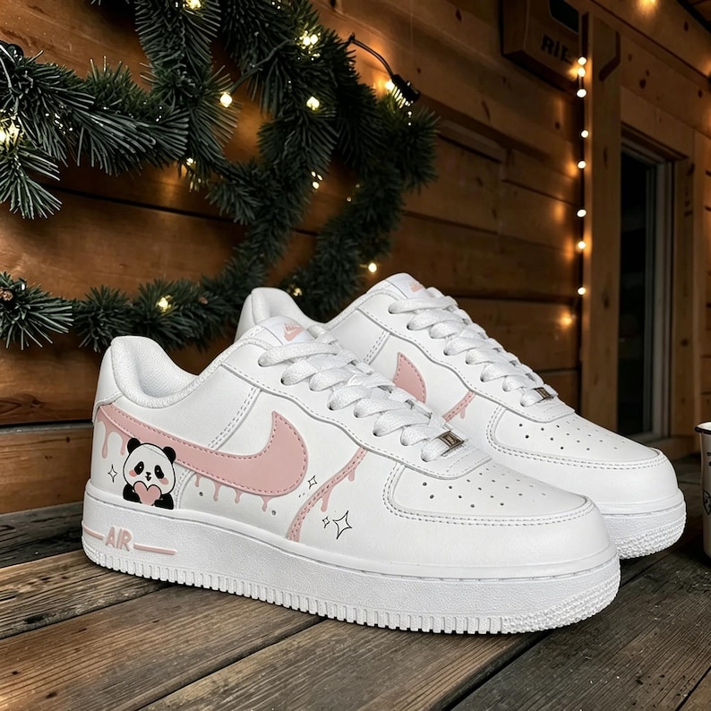 Puede incluir: Zapatillas blancas con un dise&ntilde;o de oso panda y el logo de Nike en rosa. Las zapatillas tienen una suela blanca gruesa y cordones blancos. El panda sostiene un coraz&oacute;n. La palabra "AIR" est&aacute; impresa en rosa en el lateral.