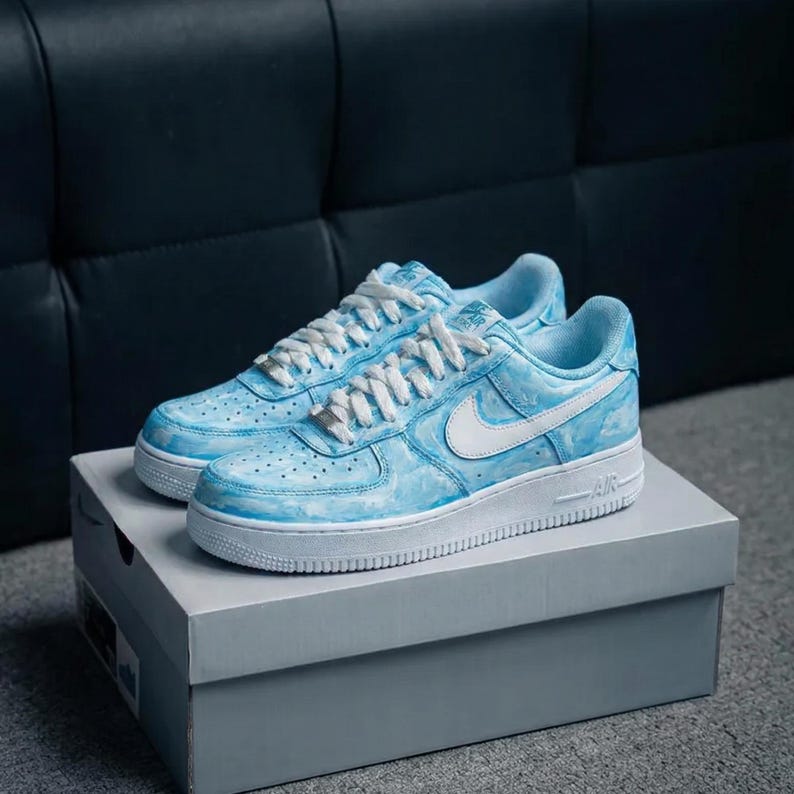 May include: A pair of light blue and white sneakers with a cloud-like design. The shoes have white laces, a white Nike swoosh, and a white sole. They are displayed on a gray box.