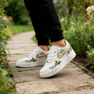 May include: White trainers with floral and gold detailing. The shoes have a gold Nike swoosh and are adorned with red, green, and gold floral patterns. They feature white laces and a white sole.