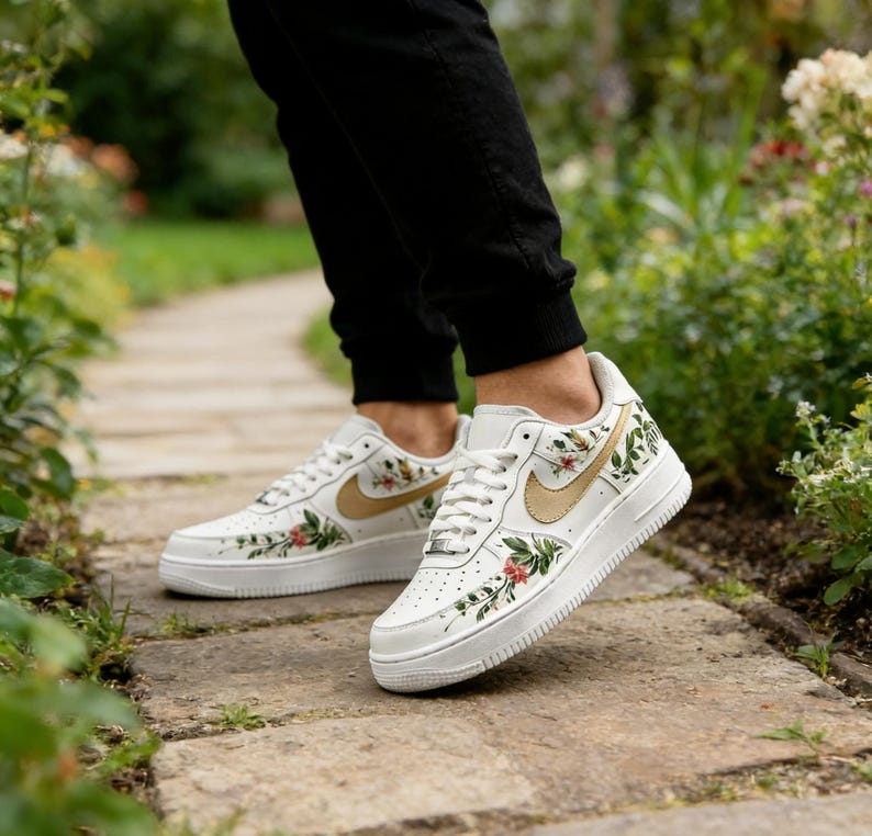 May include: White trainers with floral and gold detailing. The shoes have a gold Nike swoosh and are adorned with red, green, and gold floral patterns. They feature white laces and a white sole.