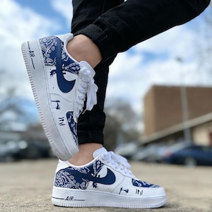 May include: White and blue Air Force 1 sneakers with a bandana pattern and navy blue swoosh. The sneakers have white laces and the word "AIR" printed on the side. The shoes are worn with black pants.