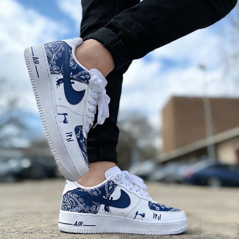 May include: White and blue Air Force 1 sneakers with a bandana pattern and navy blue swoosh. The sneakers have white laces and the word "AIR" printed on the side. The shoes are worn with black pants.