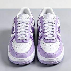 May include: Pair of white and purple Air Force 1 sneakers. The sneakers have white laces, a white sole, and a purple swoosh. The toe box and the lower part of the shoe are purple with paint splatter details.