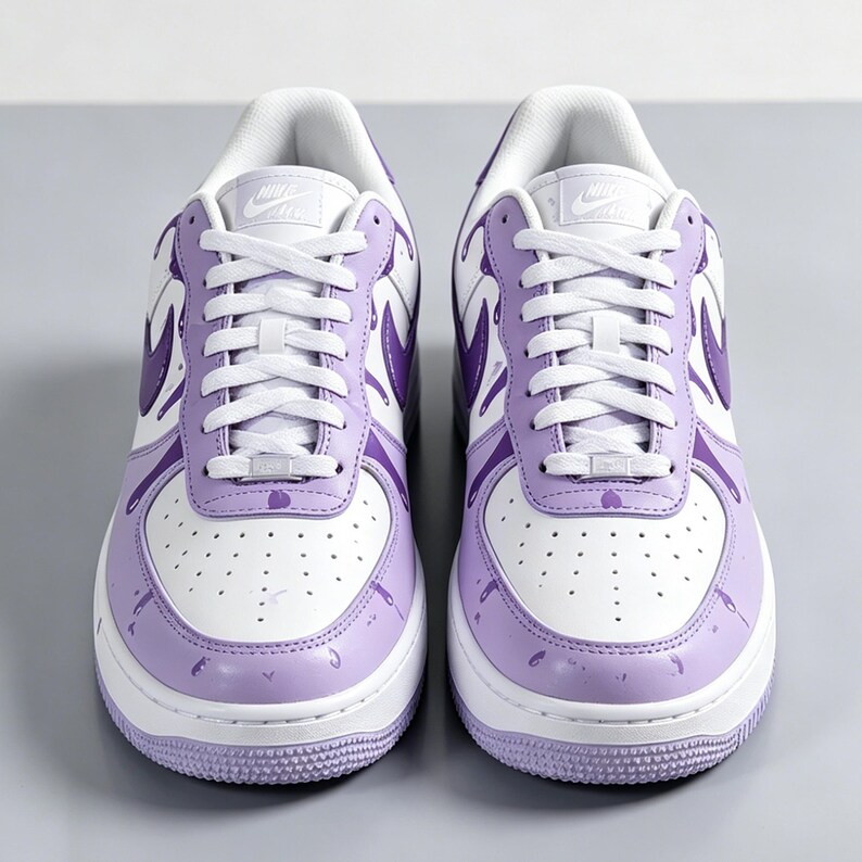 May include: Pair of white and purple Air Force 1 sneakers. The sneakers have white laces, a white sole, and a purple swoosh. The toe box and the lower part of the shoe are purple with paint splatter details.