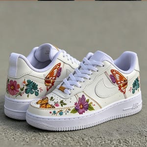May include: White sneakers with colorful floral and butterfly designs. The shoes feature a white sole, laces, and a Nike swoosh. The butterflies are orange and the flowers are pink, purple, and teal.