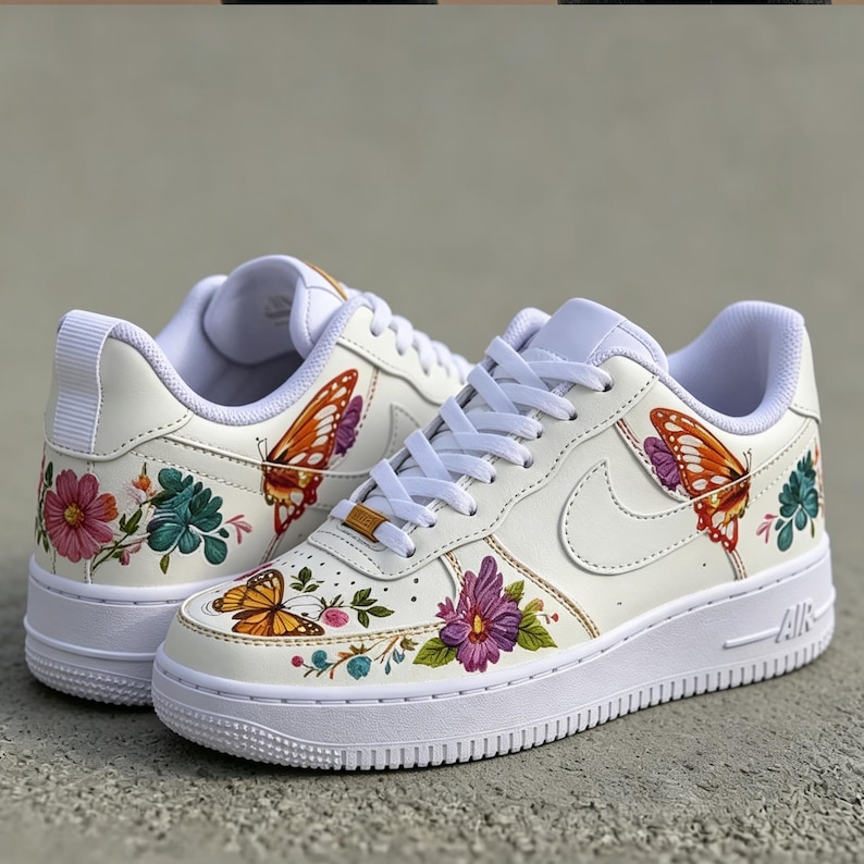 May include: White sneakers with colorful floral and butterfly designs. The shoes feature a white sole, laces, and a Nike swoosh. The butterflies are orange and the flowers are pink, purple, and teal.