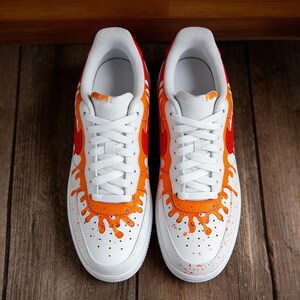May include: Pair of white sneakers with orange and red flame-like designs on the sides and heels. The shoes have white laces and soles, with orange paint splatter details. The sneakers are displayed on a wooden surface.