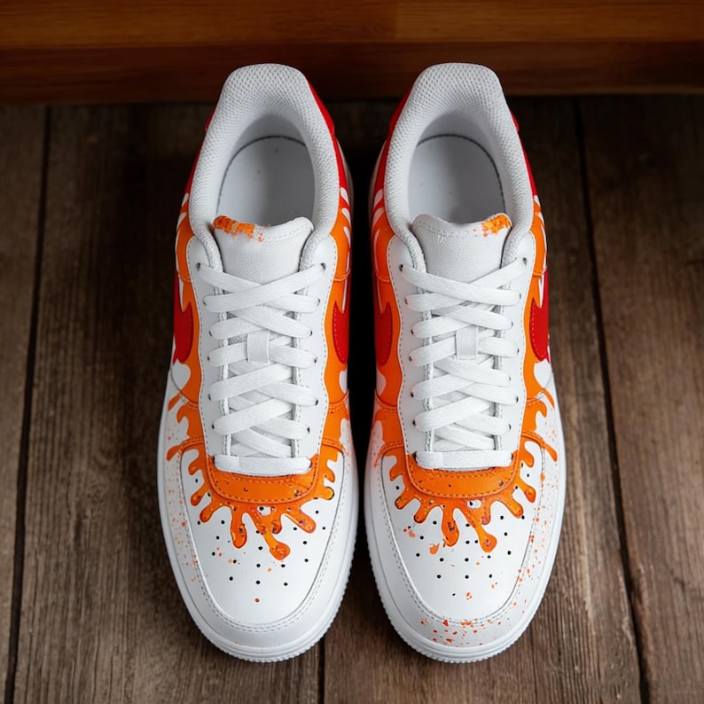 May include: Pair of white sneakers with orange and red flame-like designs on the sides and heels. The shoes have white laces and soles, with orange paint splatter details. The sneakers are displayed on a wooden surface.