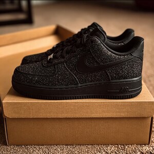 May include: A pair of black Air Force 1 sneakers with a textured, glittery upper. The shoes have a black sole, laces, and Nike swoosh. They are displayed on top of a brown cardboard box.