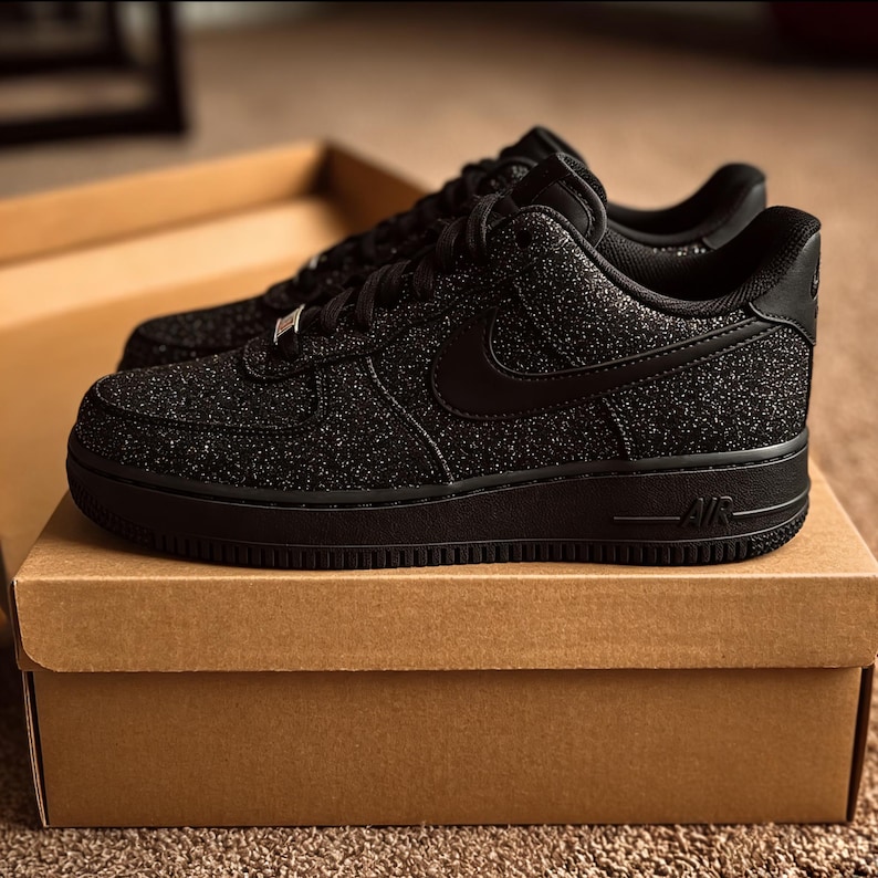 May include: A pair of black Air Force 1 sneakers with a textured, glittery upper. The shoes have a black sole, laces, and Nike swoosh. They are displayed on top of a brown cardboard box.