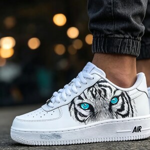 May include: White leather sneaker with a tiger eye design. The sneaker features a black and white tiger face graphic with bright blue eyes. The word "AIR" is printed on the white sole.
