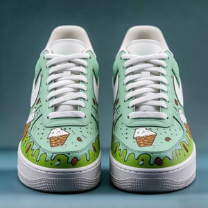 May include: A pair of mint green sneakers with white laces and soles. The sneakers are decorated with a cartoon ice cream cone design and brown almond shapes. The Nike swoosh is white.