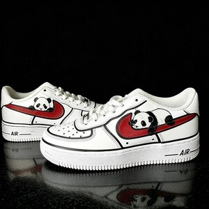 May include: White sneakers with a red swoosh and a panda bear design. The shoes have black accents and the word "AIR" on the side. The panda is lying on the red swoosh.