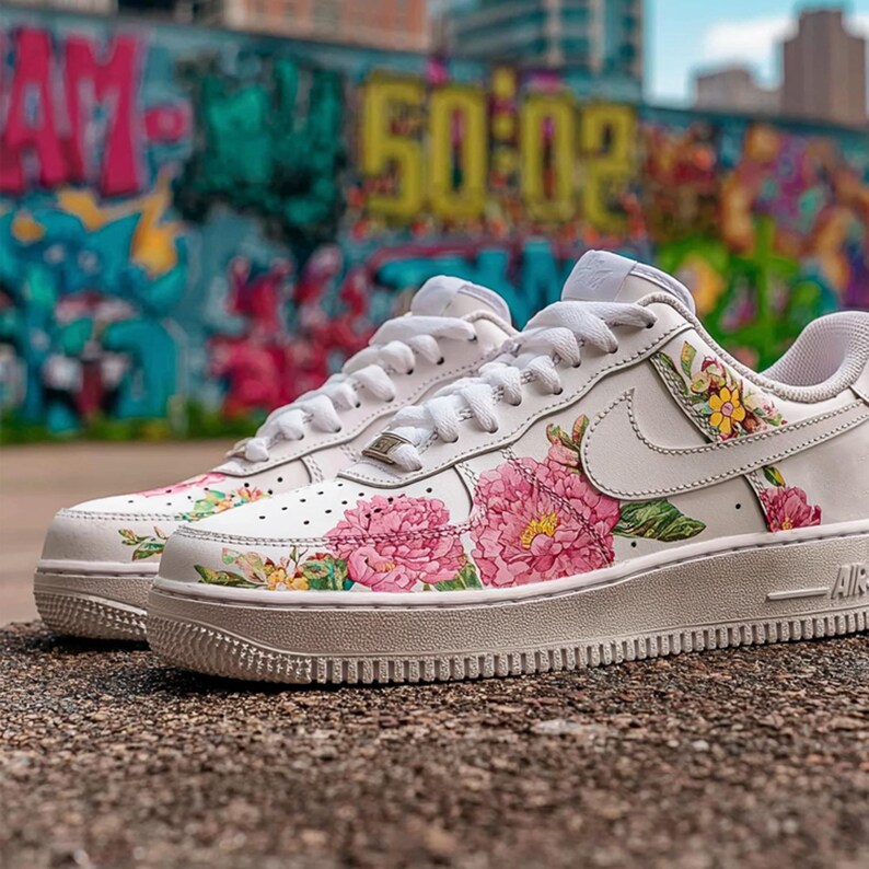 May include: White trainers adorned with a floral pattern of pink peonies and yellow flowers. The shoes have white soles and laces, with the Nike swoosh logo. The background features a vibrant graffiti mural.