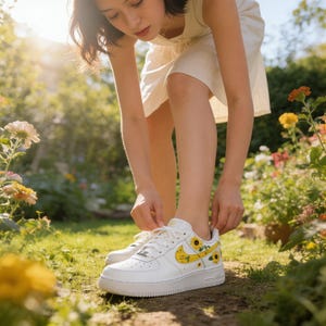 May include: White sneakers with yellow sunflower designs on the side. The sneakers have a white sole and laces. The person is tying the laces of the sneakers while standing in a garden with flowers.