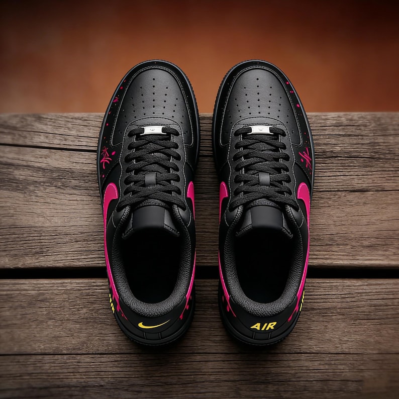 May include: Pair of black sneakers with pink accents, including the Nike swoosh logo and decorative elements. The shoes have black laces and a silver metal tag. The word "AIR" is visible on the side of one shoe. The sneakers are displayed on a wooden surface.