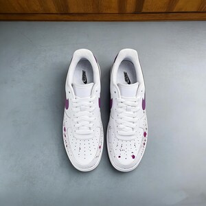 Puede incluir: Un par de zapatillas Nike Air Force 1 blancas con detalles morados. Las zapatillas presentan el logotipo de Nike morado, forros interiores y detalles de salpicaduras de pintura. Cordones y suelas blancos.