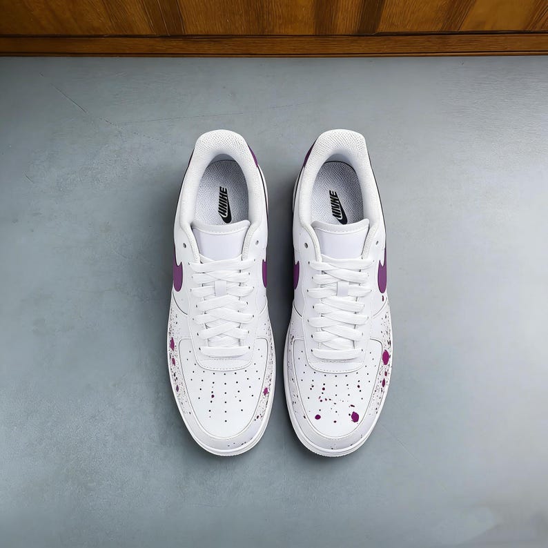 Puede incluir: Un par de zapatillas Nike Air Force 1 blancas con detalles morados. Las zapatillas presentan el logotipo de Nike morado, forros interiores y detalles de salpicaduras de pintura. Cordones y suelas blancos.