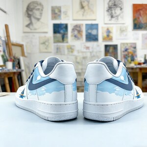 May include: White sneakers with a blue and white nautical design. The shoes feature a sailboat graphic and a sky and cloud pattern. The sneakers have a white sole and blue accents. The shoes are displayed in a studio setting with artwork in the background.