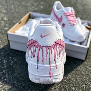 May include: White Nike Air Force 1 sneakers with pink dripping paint design. The shoes have pink laces and a pink Nike swoosh. The back of the shoe has the Nike Air logo in pink.