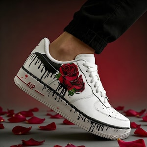 May include: White leather sneaker with a red rose design and black drip paint accents. The shoe features a red "AIR" detail and white laces. The sneaker is surrounded by red rose petals.