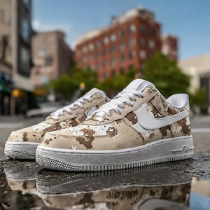 May include: Pair of tan and brown camouflage sneakers with white accents, including the iconic swoosh logo. The shoes have a white sole and laces. The sneakers are displayed outdoors on a wet surface.