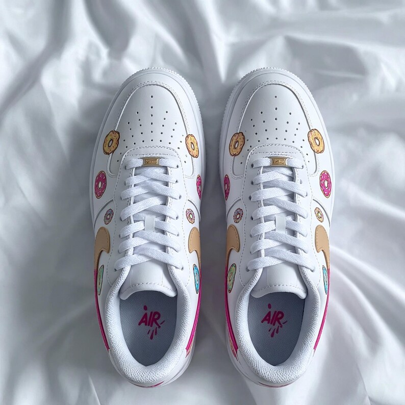 May include: Pair of white sneakers with custom donut designs. The shoes feature hand-painted donuts in various colors, including pink, yellow, and brown. The word "AIR" is printed in pink on the insole. The shoes have white laces and a white sole.