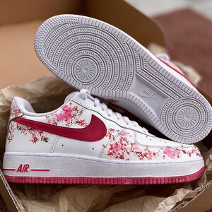 May include: White Nike Air Force 1 trainers featuring a floral design of pink blossoms and a red Nike swoosh. The sole is white with a red trim. The word "AIR" is printed in red on the side.