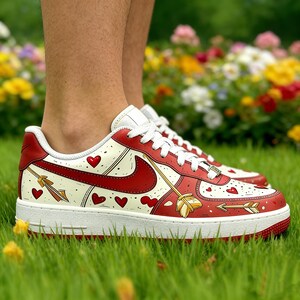 May include: Red and white sneakers with heart and arrow designs. The shoes feature a red Nike swoosh, red accents, and a white sole. The design includes red hearts and gold arrows, creating a romantic theme.
