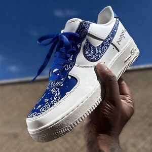 May include: White and blue Air Force 1 sneakers with a custom design. The shoe features a blue paisley pattern on the toe, heel, and Nike swoosh. The laces are blue, and the word "Angus" is written on the side.