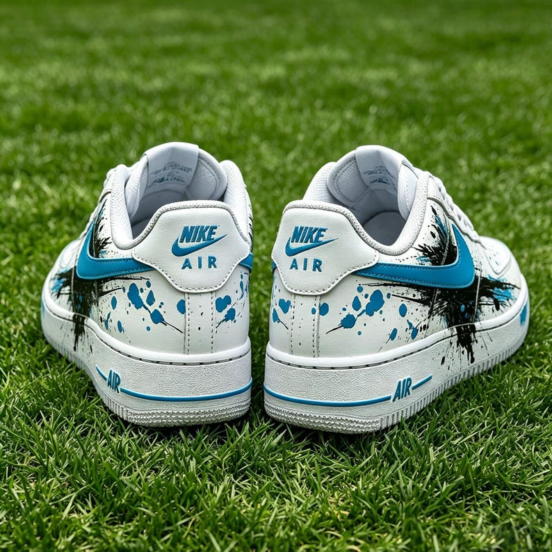 May include: White Nike Air Force 1 sneakers with blue and black paint splatter design. The sneakers have a white sole with blue accents and the Nike Air logo on the heel. The shoes are on a green grass background.