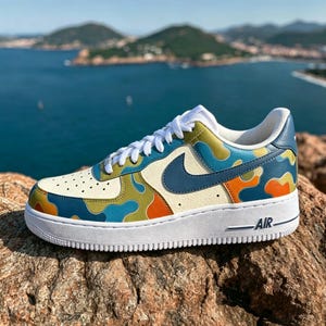May include: A custom-painted Air Force 1 sneaker with a white sole and a cream-colored upper. The shoe features a blue Nike swoosh and a camouflage pattern in blue, green, and orange. The word "AIR" is printed on the side of the white sole.