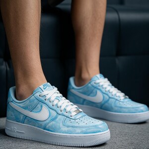 May include: Light blue Nike Air Force 1 sneakers with a marbled design and white accents. The sneakers feature white laces, a white Nike swoosh, and a white sole. The word "AIR" is visible on the side of the sole.