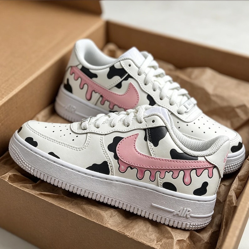 Op de afbeelding: Witte sneakers met een zwart-roze koeienprint ontwerp. De schoenen hebben een roze Nike swoosh en een witte zool. De sneakers liggen in een bruine kartonnen doos.