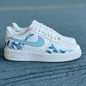 May include: White sneakers with light blue floral design accents. The sneakers feature a light blue Nike swoosh and floral patterns on the sides. The soles are white, and the word "AIR" is visible on the side of the sole.