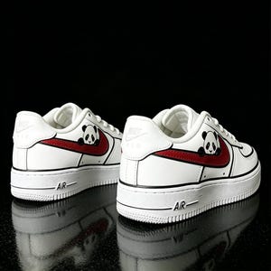 May include: White sneakers with a red swoosh and a panda bear graphic. The shoes have a thick white sole and the word "AIR" is printed on the side. The Nike logo is visible on the heel.