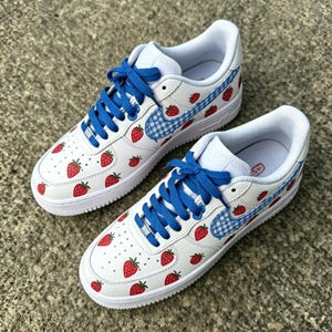 May include: White sneakers with red strawberry graphics and blue laces. The Nike swoosh is a blue and white gingham pattern. The shoes have a white sole and a white interior.