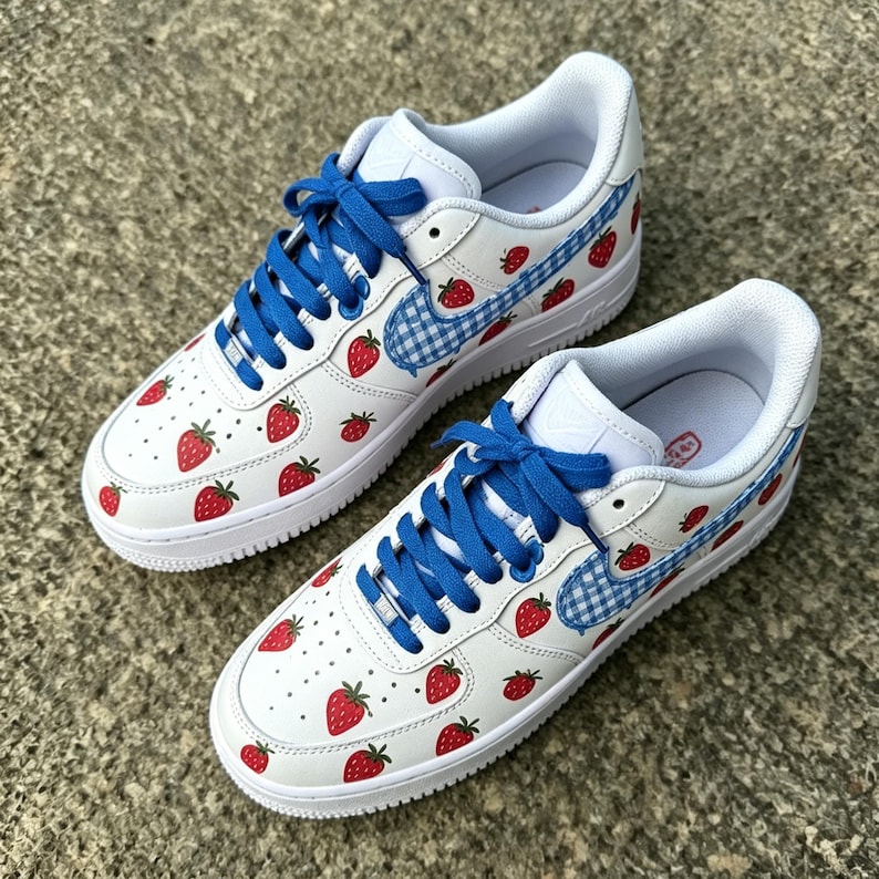 May include: White sneakers with red strawberry graphics and blue laces. The Nike swoosh is a blue and white gingham pattern. The shoes have a white sole and a white interior.