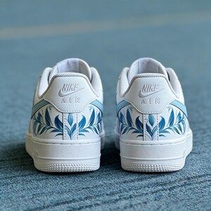 May include: Pair of white Nike Air Force 1 sneakers with light blue accents and floral leaf design. The sneakers feature the Nike Air logo on the heel and a white sole. The design is a unique take on a classic shoe.
