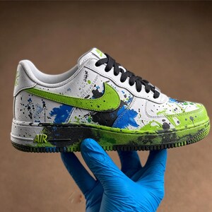 May include: White and green Air Force 1 sneakers with black laces. The shoes feature a green Nike swoosh and are decorated with blue, green, and black paint splatters. The sole is green and black.