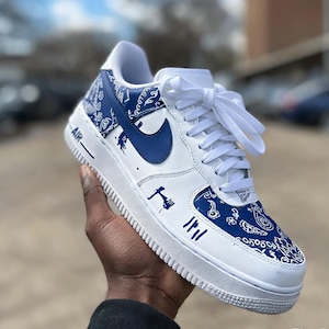 May include: White and blue Air Force 1 sneakers with a paisley pattern on the toe and heel. The Nike swoosh is blue. The shoe has white laces and the word "AIR" is printed on the side.