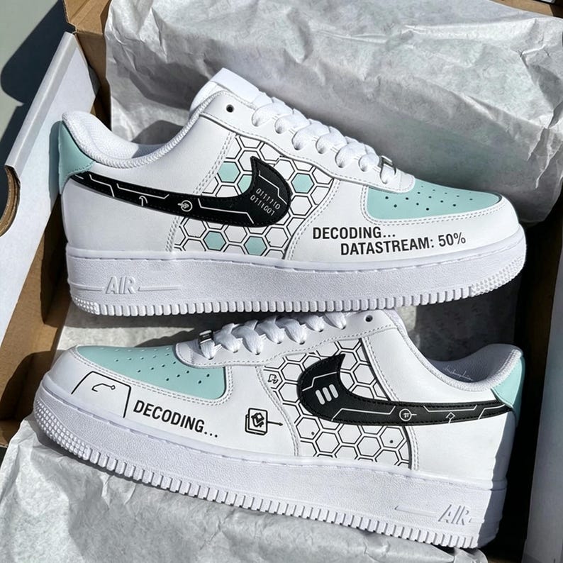 May include: White sneakers with a light blue toe and accents. The shoes feature black graphics, including a honeycomb pattern and the text "DECODING... DATASTREAM: 50%." The shoes have a white sole and the word "AIR" on the side.