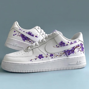 May include: White sneakers with purple floral branch designs. The shoes feature a classic design with a thick white sole and the word "AIR" on the side. The purple flowers and branches are painted on the sides and back of the shoes.