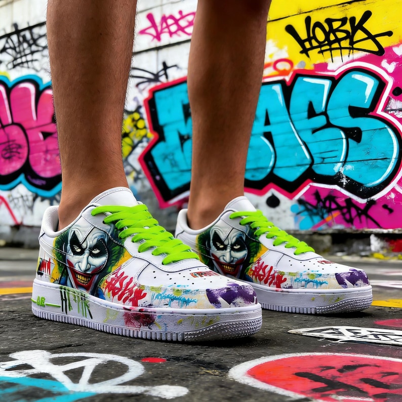 May include: White sneakers with a Joker-themed design. The shoes feature a painted image of the Joker on the sides, with green laces and colorful graffiti-style accents. The soles are white with colorful paint splatters.