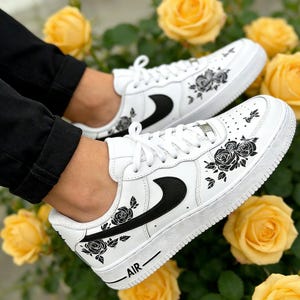 May include: White sneakers with black rose floral designs and a black swoosh logo. The word "AIR" is printed on the side of the shoe. The shoes are laced with white laces.